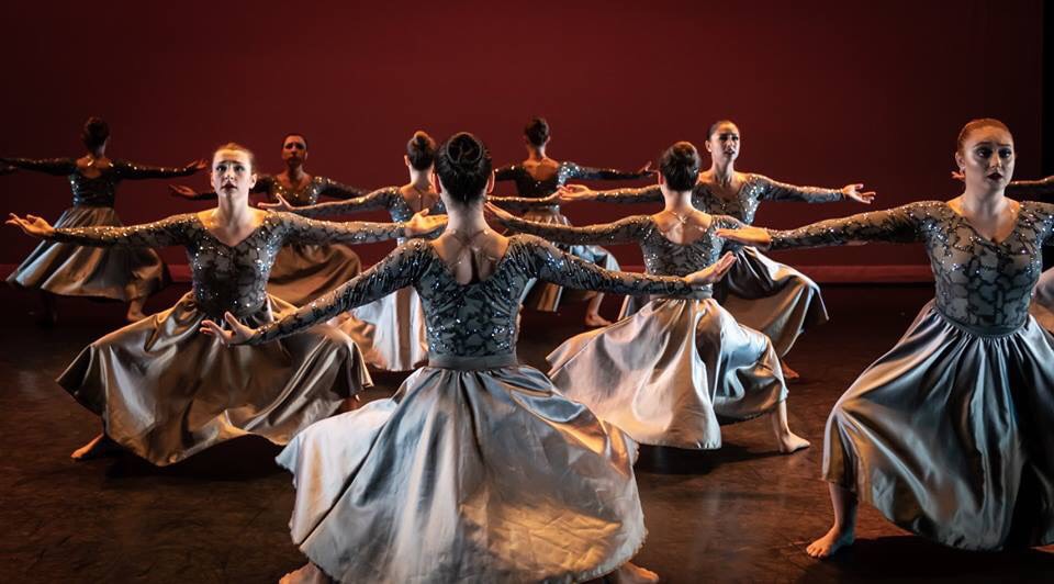 Dance ensemble performing in silver costumes on a theater stage.
