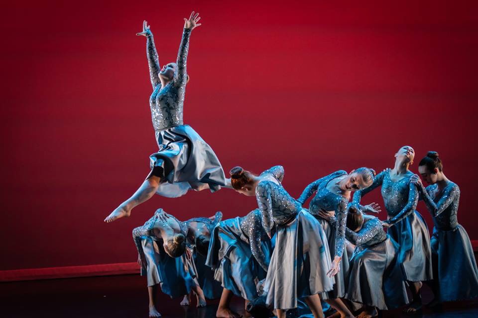 Dancer leaping above an ensemble against a red stage backdrop.